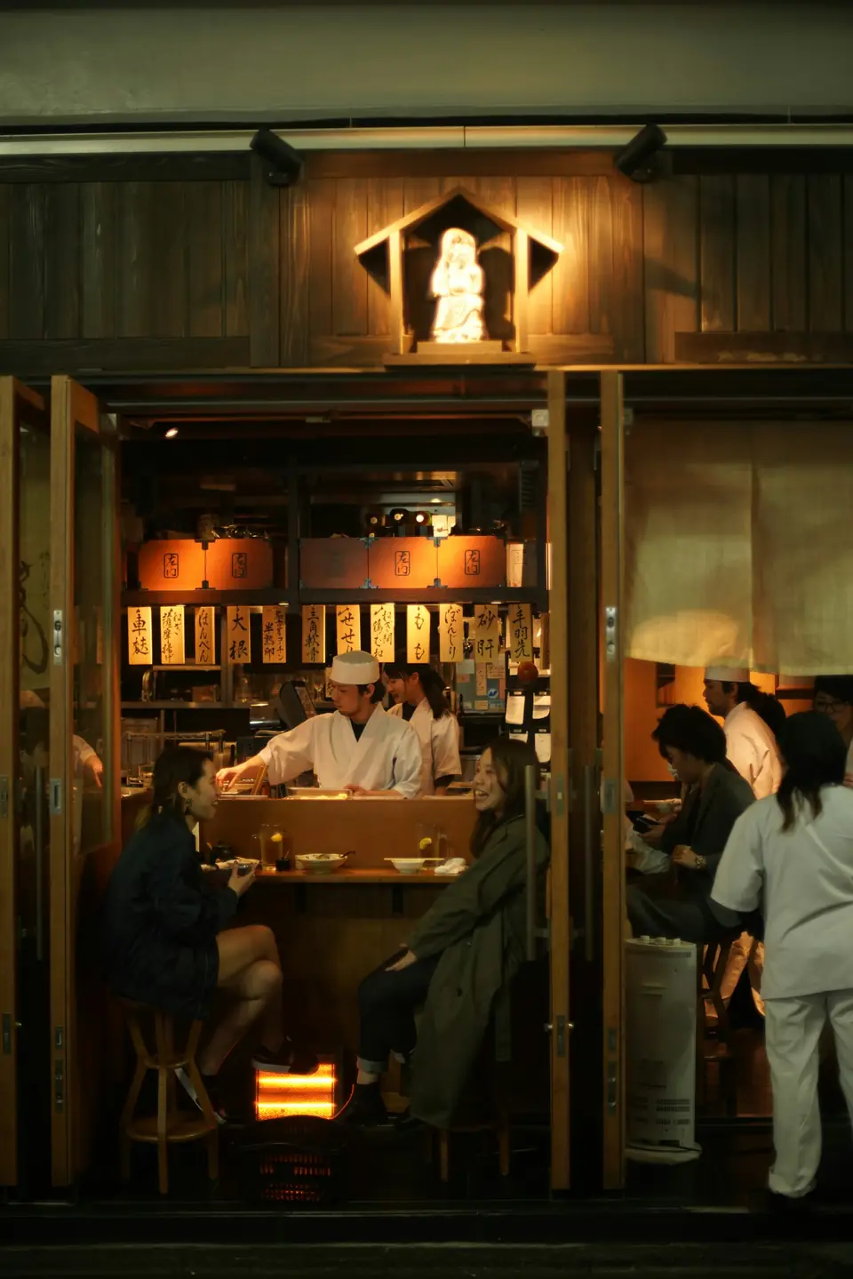 A warm izakaya counter with a chef and guests seen through the doorway
